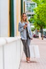 Frontansicht einer positiven Frau im schicken Anzug mit Coffee to go, die in die Kamera schaut, während sie ein Paar Einkaufstüten in der Hand hält — Stockfoto