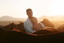 Hombre abrazando tierna mujer con los ojos cerrados de pie cerca entre caballos tranquilos en el campo montañoso en la luz del atardecer - foto de stock