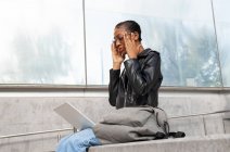 From below African American female freelancer in leather jacket sitting with netbook on laps during online work on street while he puts on his glasses — Stock Photo