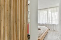Interior del dormitorio contemporáneo con cama entre tabique de madera y ventana con tul en casa de luz - foto de stock