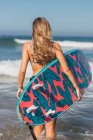 Vista posterior de una surfista deportiva anónima con tabla de surf paseando en el mar ondulado durante el entrenamiento en un resort tropical en un día soleado - foto de stock