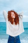 Восхитительная женщина с кудрявыми длинными рыжими волосами в трикотажном свитере, стоящая с руками за головой и закрытыми глазами против синего моря — стоковое фото