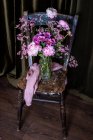 Buquê de peônias coloridas frescas e crisântemos em vaso de vidro colocado em cadeira de madeira desgastada perto de cortinas na sala de luz — Fotografia de Stock