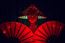 Donna irriconoscibile con una maschera in abito tradizionale creativo e copricapo vietnamita con illuminazione rossa in piedi in studio scuro su sfondo nero durante la performance — Foto stock
