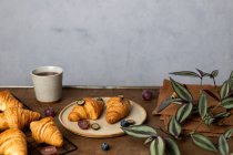 Вкусные свежие круассаны подаются на тарелке с фруктами, помещенными возле чашки чая на деревянном столе утром в светлом помещении — стоковое фото