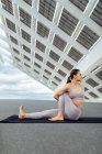 Mujer deportiva de cuerpo completo en ropa deportiva sentada en la esterilla mientras practica yoga asana con giro cerca del panel solar en la calle - foto de stock
