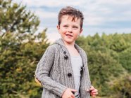 Adorable petit garçon en élégant cardigan tricoté chaud souriant et regardant loin tout en se tenant contre des arbres verts luxuriants contre un ciel nuageux dans la campagne — Photo de stock