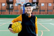 Mujer madura optimista en ropa deportiva y auriculares mirando a la cámara mientras está de pie en la cancha pública de baloncesto con pelota durante el entrenamiento - foto de stock
