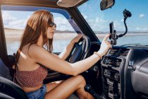 Side view of confident young female traveler with long ginger hair hair in summer clothes using navigator on mobile phone while driving modern car near sea on sunny day — Stock Photo