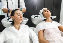 Giovani donne in vestiti bianchi chiudono gli occhi mentre si lavano i capelli nel moderno salone di bellezza — Foto stock