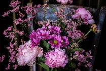 Bouquet di peonie fresche colorate e crisantemi in vaso di vetro posto su sedia in legno intemperie vicino a tende in stanza luminosa — Foto stock
