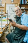 Концентрований старший майстер чоловічої статі в фартухах та окулярах, використовуючи мобільний телефон, сидячи на верстаті під час друку в студії — стокове фото