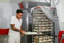 Jeune boulanger masculin tatoué élégant dans des vêtements décontractés et des lunettes mettant des pains de pâte crue sur un support métallique pendant la cuisson du pain dans la cuisine — Photo de stock