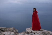 Dreamy female with long hair in Victorian styled outfit with cape standing above endless sea against cloudy sky in nature — Stock Photo