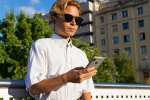 Homem jovem confiante em mensagens de texto camisa branca no celular, enquanto em pé na rua contra o céu azul no dia ensolarado — Fotografia de Stock
