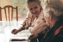 Sœurs âgées inspectant des photos dans un album photo et discutant de souvenirs assis à table à la maison ensemble — Photo de stock