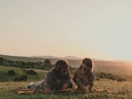 Hippie couple with sunglasses interacting while lying on grassy meadow in nature at sunset time — Stock Photo