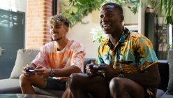 Веселі багаторасові друзі з геймпадами в руках сидять на дивані, граючи в відеогру разом у світлій вітальні з зеленою рослиною — стокове фото