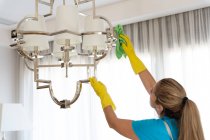 Vista lateral de una joven limpiadora profesional femenina con pelo largo en uniforme y guantes limpiando la lámpara con paño de polvo durante el trabajo en un elegante apartamento - foto de stock