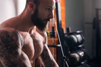 Vista lateral del deportista muscular concentrado con tatuajes haciendo ejercicios en la máquina de cruce de cables en el gimnasio con paredes ligeras - foto de stock
