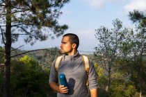 Pensive young bearded Hispanic male hiker in casual clothes standing in lush green forest with thermos in hand and looking away on sunny day — Stock Photo