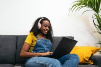 Femme noire souriante en salopette denim avec écouteurs assis sur le canapé et utilisant un ordinateur portable à la maison — Photo de stock