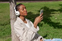 Спокойная афроамериканка с закрытыми глазами, слушающая музыку в беспроводных наушниках, сидя на газоне возле ствола дерева в солнечном парке, используя смартфон — стоковое фото