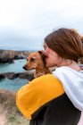Счастливая женщина-путешественница целует милого щенка, стоя на лугу в высокогорье в playa de las catedrales в Галисии на закате — стоковое фото