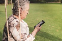 Seitenansicht einer Seniorin mit Bluse, die im Park sitzt und elektronisches Buch liest — Stockfoto