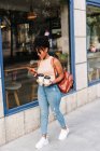 Full body of trendy young ethnic female millennial with dark Afro hair in stylish outfit and backpack using mobile phone while walking on city street with cups of takeaway coffee — Stock Photo