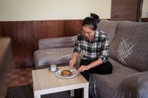 Молодая азиатская женщина ест домашние блинчики на тарелке возле чашки кофе на столе, сидя на удобном диване в гостиной — стоковое фото