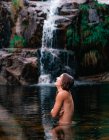 Vue latérale de sourire femme nue voyageant couvrant poitrine et debout dans l'eau calme de l'étang près de la cascade dans le parc naturel de Fervenza De Casarinos en Espagne — Photo de stock
