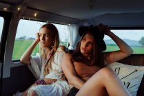 Duas lindas garotas caucasianas em roupas de verão deitadas no assento dentro de uma van vintage olhando para longe — Fotografia de Stock