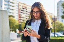 Dal basso di focalizzata donna d'affari in abiti alla moda e occhiali in piedi e smartphone di navigazione contro alberi verdi nel parco in estate alla luce del giorno — Foto stock