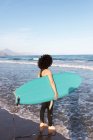 Back view of unrecognizable female surfer in wetsuit with surfboard standing looking away on seashore washed by waving sea — Stock Photo