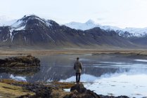 Rückansicht eines unkenntlich gemachten männlichen Touristen in Freizeitkleidung, der die wilde Natur bewundert, während er in der Nähe eines friedlichen Sees steht, der die schneebedeckten Berge Islands reflektiert — Stockfoto