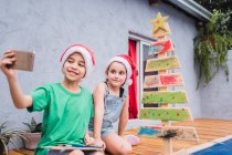 Crianças otimistas tomando auto-retrato no smartphone enquanto se sentam perto da árvore de Natal de madeira no quarto durante a preparação de férias juntos em casa — Fotografia de Stock