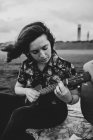 Talentoso músico feminino com cabelo castanho em roupas casuais tocando ukulele e cantando música enquanto sentado na praia de areia na natureza à luz do dia — Fotografia de Stock