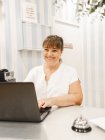 Administrador feminino adulto sorridente em uniforme branco olhando para a câmera enquanto trabalhava no laptop na mesa perto do sino no salão de beleza — Fotografia de Stock
