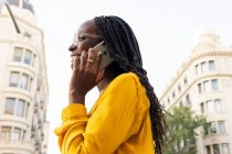 Femme afro-américaine positive dans des lunettes regardant loin tout en ayant une conversation téléphonique dans la rue avec des bâtiments résidentiels et des arbres en ville — Photo de stock