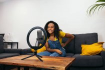 Sonriente hembra negra en el sofá mientras usa el teléfono inteligente en la lámpara de anillo led cerca de luces profesionales en trípodes - foto de stock