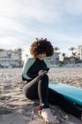 Позитивная женщина с вьющимися волосами в гидрокостюме, просматривающая смартфон, сидя на песчаном пляже с доской для серфинга — стоковое фото