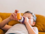 Вид сбоку маленького мальчика с астмой, использующего ингалятор, лежа на диване дома — стоковое фото