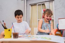 Positive kids with paintbrushes painting with colorful watercolors on paper at table with with supplies in light room with whiteboard — Stock Photo