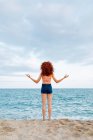Вид ззаду на невпізнавану жінку з кучерявим рудим волоссям, що робить жест дзен на березі блакитного розривного моря — стокове фото