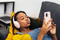 Von oben eine junge Afroamerikanerin mit lockigem Haar, Kopfhörer und lässiger Kleidung, die auf einem bequemen grauen Sofa liegt und im hellen Zimmer zu Hause ein Selfie mit dem Smartphone macht — Stockfoto