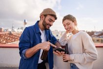 Smiling bearded man with bottle of beer hugging positive girlfriend scrolling mobile phone on balcony in sunny day — Stock Photo