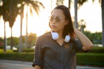 Mujer asiática seria con auriculares blancos modernos mirando a la distancia mientras está de pie cerca de la carretera en la calle de la ciudad con árboles verdes - foto de stock