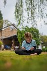Corpo inteiro de menina descalça concentrada lendo livro interessante enquanto sentado no gramado gramado no quintal contra edifício residencial no campo — Fotografia de Stock