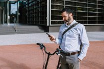 Maschio manager a piedi con bicicletta e lettura di messaggi su smartphone nel parco — Foto stock